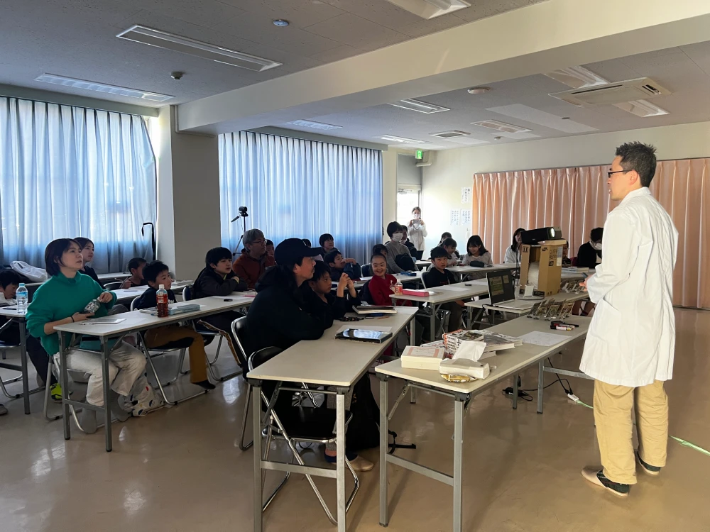 白衣姿のよーた先生がスクリーンの前で子どもたちに昆虫の話をするようす